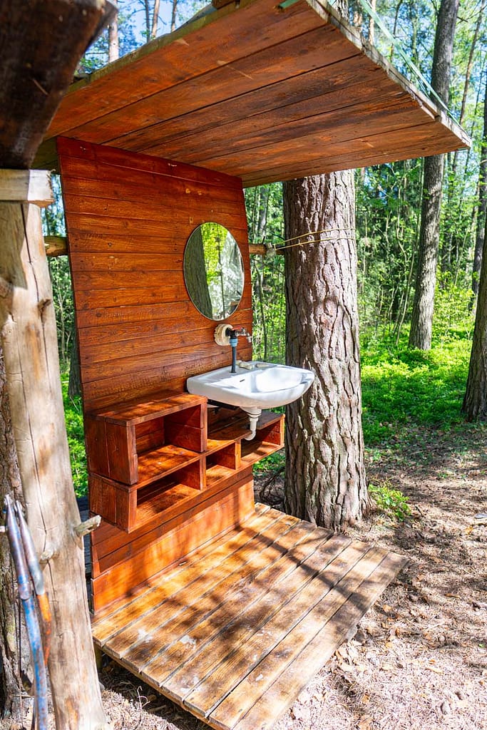 Venkovní koupelna s policemi, umyvadlem a zrcadlem , umístěné mezi stromy v přírodním prostředí před jurou Rozárkou.
