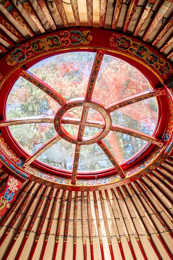 Detailní pohled na střešní okno jurty Rozárky - dřevěná konstrukce s červenými trámy a kruhovým světlíkem, kterým proudí denní světlo dovnitř.