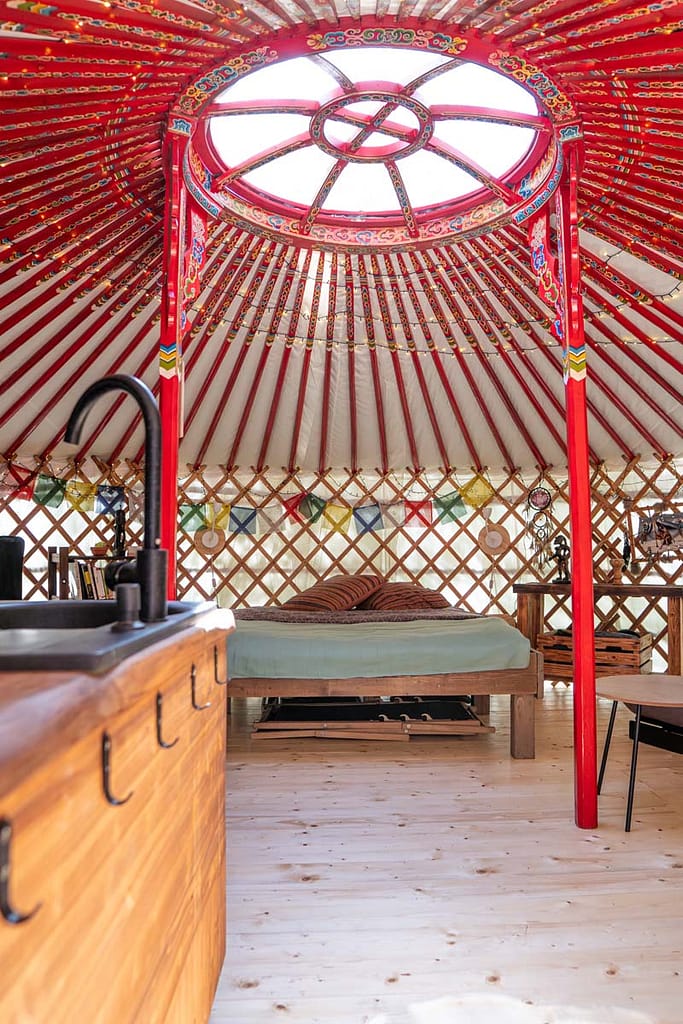 Pohled do interiéru jurty Rozárky s červenými středovými sloupy, kuchyňským koutem a pohodlnou postelí pod střešním světlíkem, který propouští denní světlo.