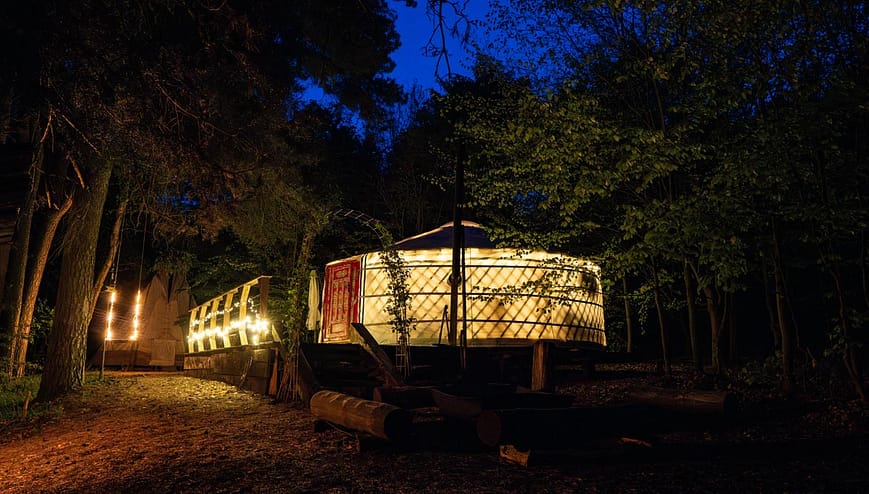 Noční pohled na jurtu Rozárku osvětlenou teplým světlem a řetězem světýlek. atmosférické prostředí uprostřed lesa s posezením na terase.
