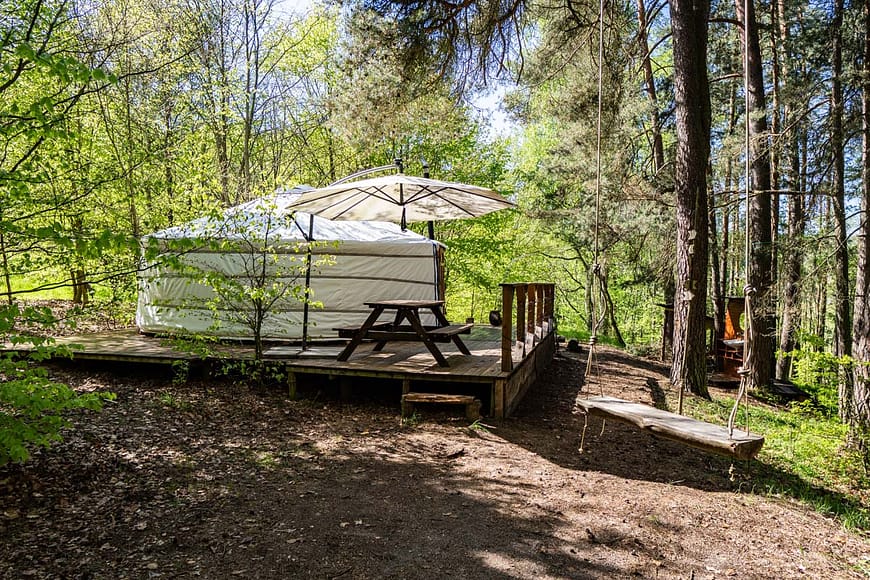 Jurta Rozárka umístěna na dřevěné terase uprostřed lesa. Před jurtou Rozárkou je posezení se slunečníkem, obklopené stromy a přírodními prvky.