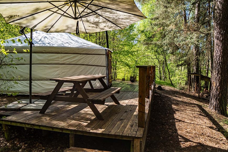 Jurta Rozárka umístěna na dřevěné terase uprostřed lesa. Před jurtou Rozárkou je posezení se slunečníkem, obklopené stromy a přírodními prvky.